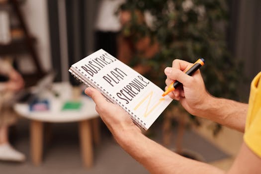 Close-up of a hand writing in a notebook with 'Business Plan Schedule' indoors.